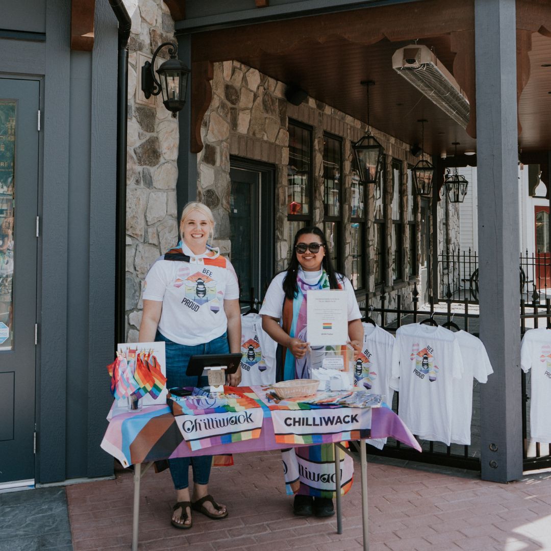 Two people stand behind a table at the Pride Festival, with various pride merchandise on display.