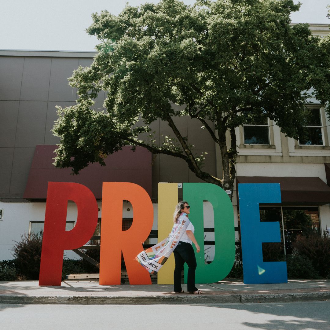 A person wearing a pride flag as a cape stands in front of a rainbow-coloured pride sign.