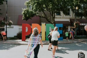 A woman wearing a pride flag as a cape walks up to a large, rainbow coloured pride sign in Downtown Chilliwack.