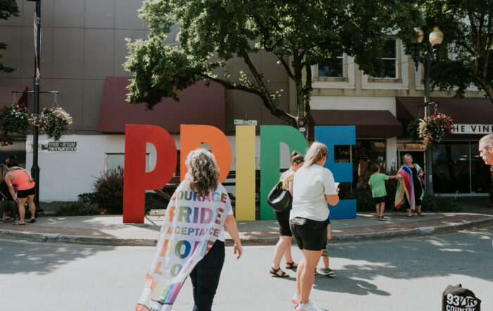 A woman wearing a pride flag as a cape walks up to a large, rainbow coloured pride sign in Downtown Chilliwack.