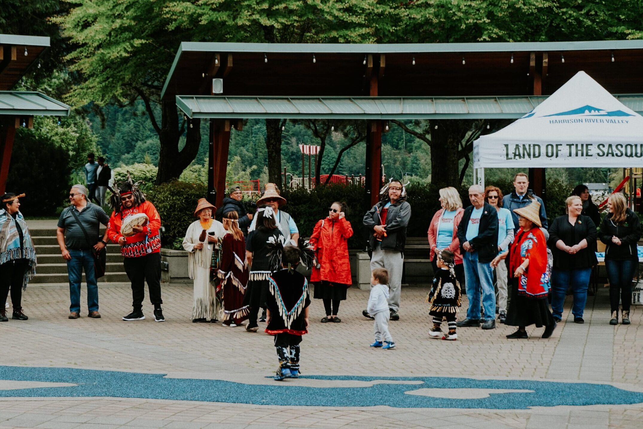 A group of people stand in a plaza, some wearing Indigenous regalia.