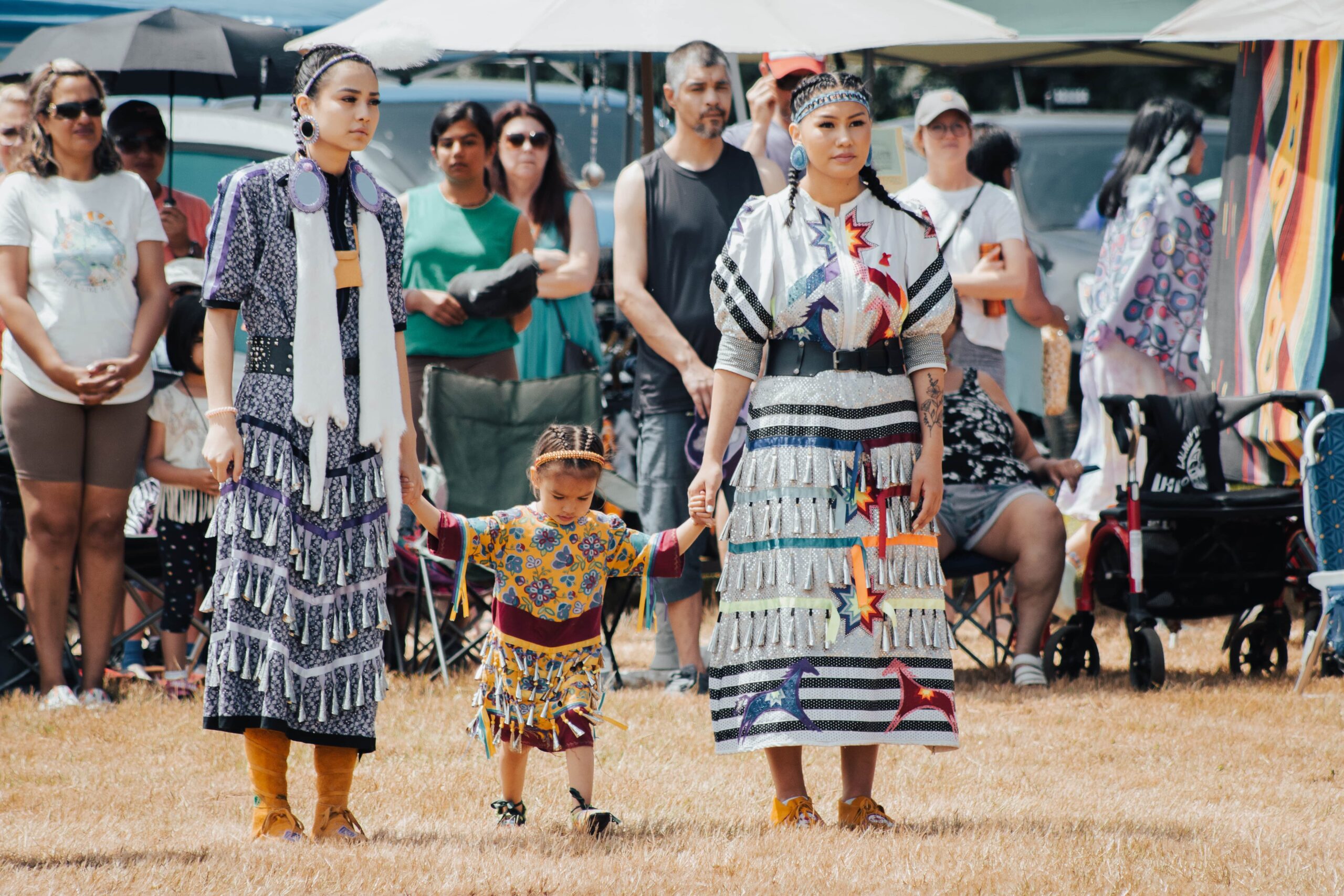 Two women stand holding the hands of a young child in-between them, all wearing traditional Indigenous regalia.