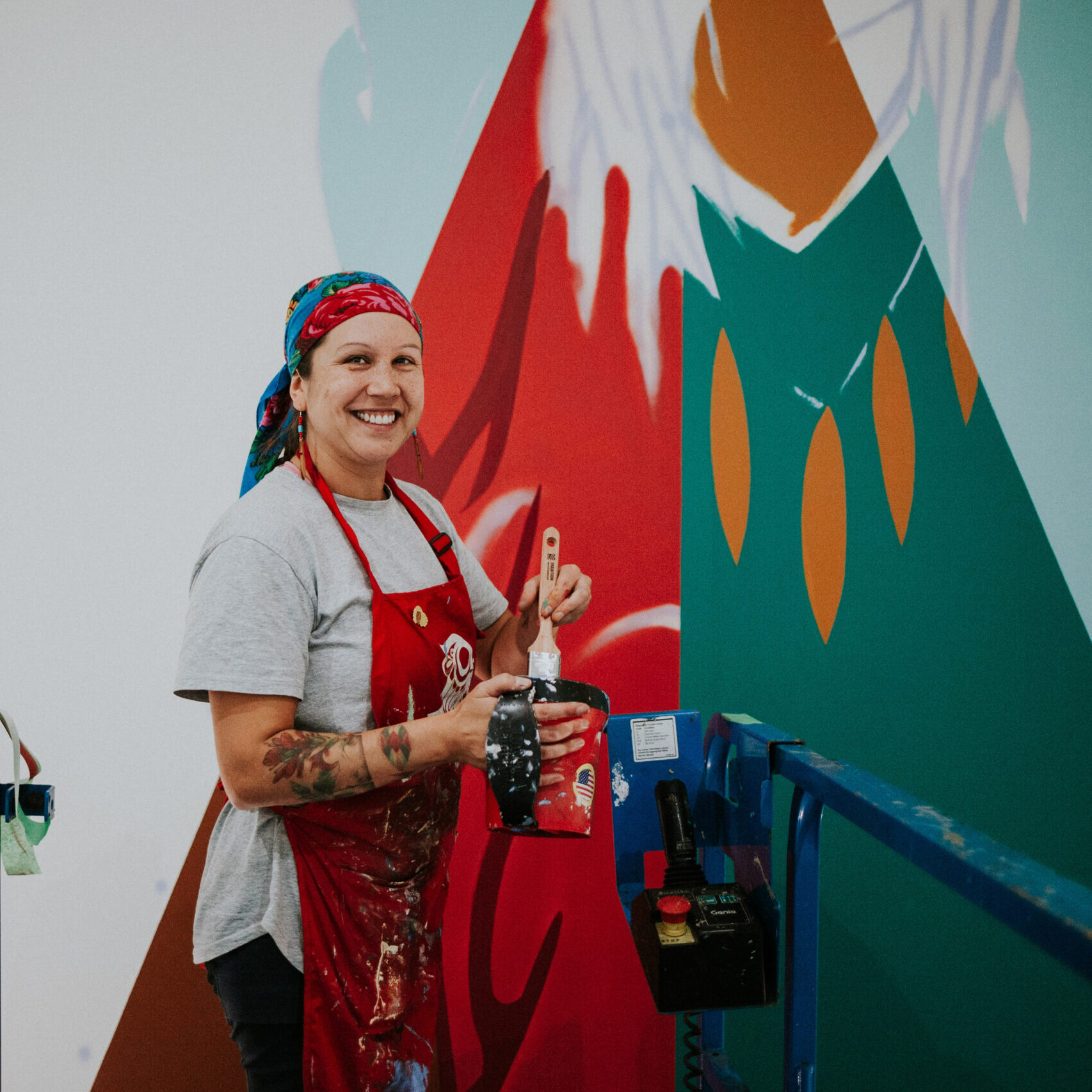 A woman stands in front of an Indigenous art mural that she is currently painting.